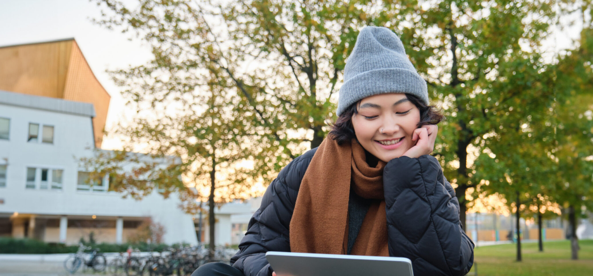 Portrait of asian girl relaxing in park, watching videos or reading on digital tablet, sitting on bench in hat and scarf on chilly day, enjoing outdoors.